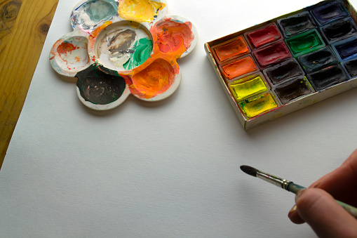 A woman paints with watercolors. Hand with a brush, watercolor paints and a palette on a background of white paper and a table.