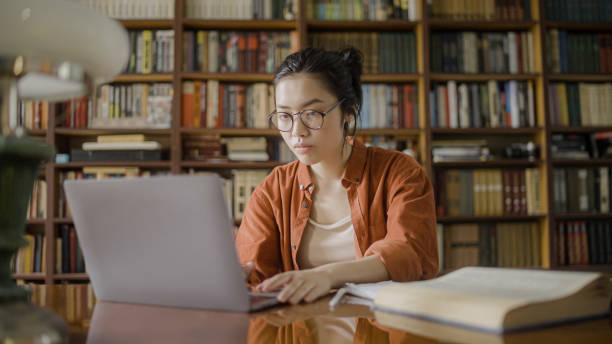 istock Focused young woman doing research in the library, typing an article on laptop 1455207788