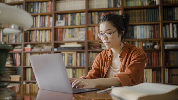 istock Focused young woman student spending time in university library, doing research, typing on laptop 1455207856