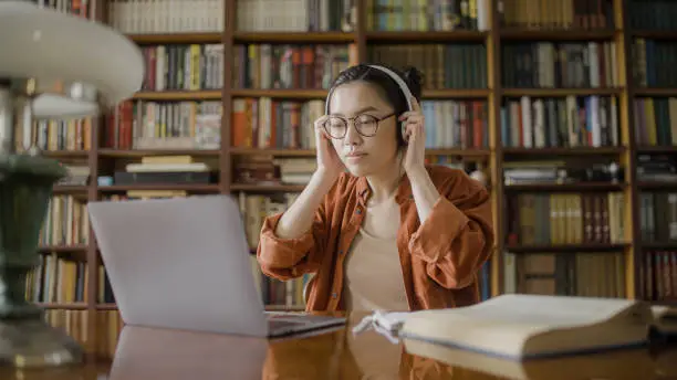 Young asian woman putting on headphones, making video call on laptop in library