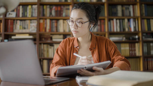 istock Young woman student taking online course, sitting with laptop in the library 1455208312