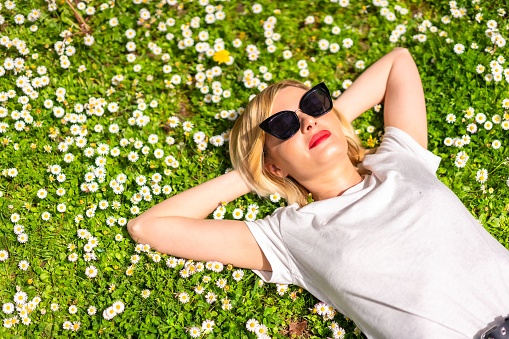 A closeup shot of a young blonde girl in a hat and sunglasses lying on the grass and relaxing in the park