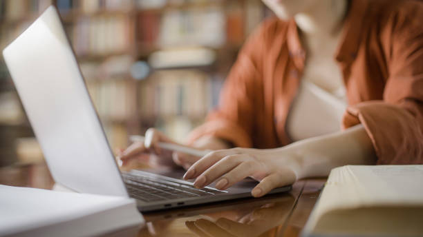 Female student doing research in a library, typing on laptop, reading books