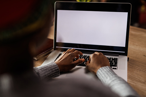 Over the shoulder view of young diligent woman sitting at her desk at the office and typing data on a laptop with blank, white screen.