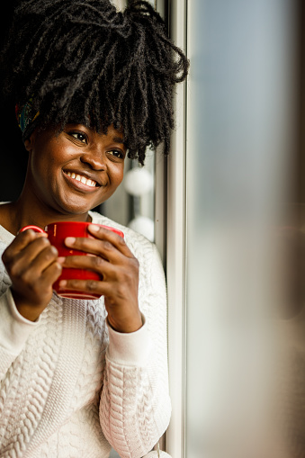 Portrait of joyful young woman sitting by the window, enjoying a cup of tea or coffee, looking outside, smiling and contemplating about Christmas.