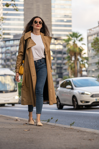 Adult 35s years old businesswoman with long hair, sunglasses and trench coat walk in a town with modern buildings. Urban scene of Barcelona city.