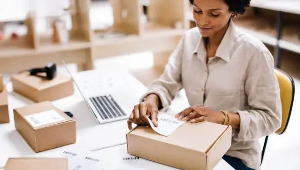 Female entrepreneur sticking a barcode label onto a package box. Online store owner preparing an order for shipping in a warehouse. Creative businesswoman running an e-commerce small business.