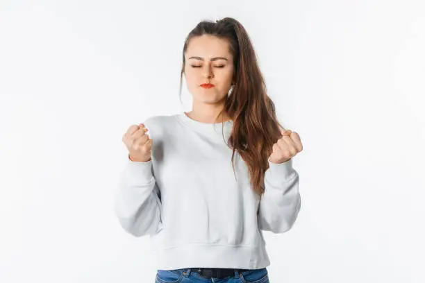 Pissed-off girl gesticulating with her hands, stands over white background with close eyes. Distressed and irritated female in white sweatshirt squeeze hands in fists losing patience, boiling from annoyance