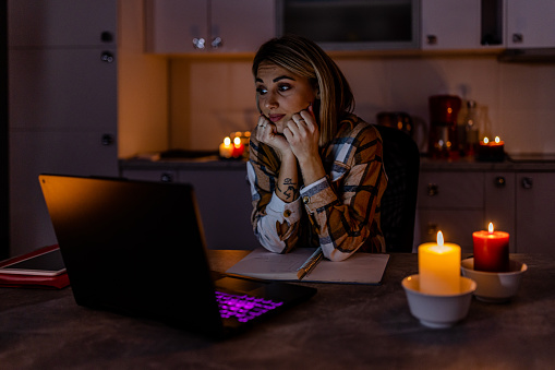 In the time of a blackout caused by an energetic crisis, a woman is working on a laptop at home under lit candles.