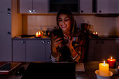 During an energetic crisis, a woman is working on a laptop using a mobile phone in the dark with lit candles.