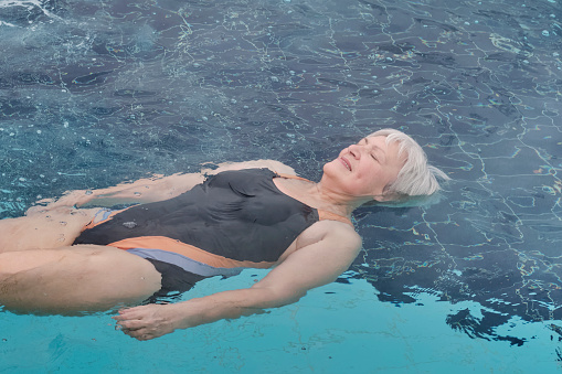 Mature woman with short gray hair, eyes closed, in swimsuit lying on back on water, relaxing. Outdoor thermal pool. Mental health concept