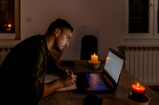 During an energetic crisis, a man is working on a laptop in the dark with lit candles.