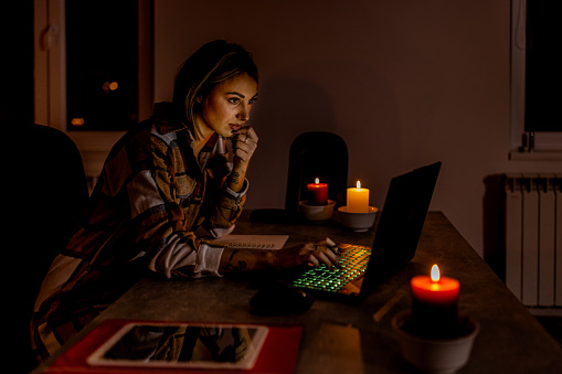 A young woman is working during an electricity crisis causing a blackout under the lit candles using a laptop.