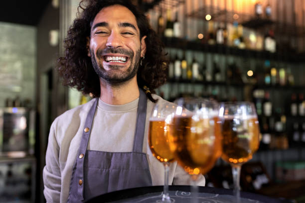 garçom jovem sorridente feliz vestindo avental em pé no bar segurando bandeja de cervejas olhando para a câmera. - beer glass mustache beer color image - fotografias e filmes do acervo