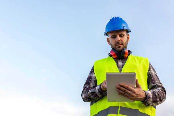 istock Man wearing helmet and vest checking information on digital tablet outdoors. Blue sky, copy space. 1455226650