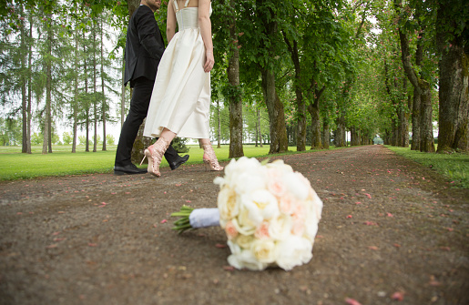 Wedding bouquet of white roses lying on the ground after the wedding.  Bride and groom leaving on treelined path.