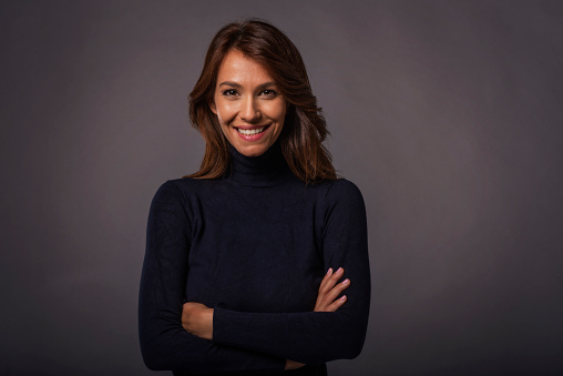 Close-up of an attractive middle aged woman with toothy smile wearing black turtleneck sweater while staning at isolated dark background. Copy space. Studio shot.