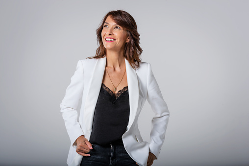 Attractive middle aged woman with toothy smile wearing blazer while standing at isolated dark background. Copy space. Studio shot.