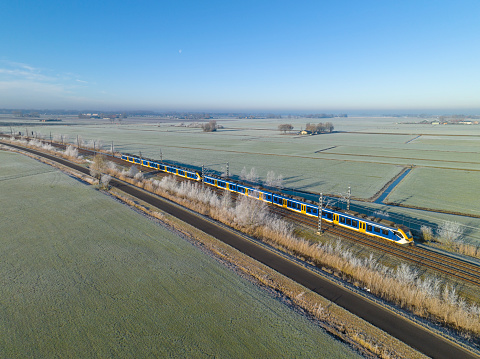 Commuter train of the Nederlandse Spoorwegen (NS) driving through the countryside landscape on the Hanzelijn in The Netherlands seen from above during a cold winter morning. The landscape is cold and frozen during an early winter morning.
