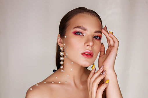 Portrait of young female model with makeup bare shoulders and pearls on face looking at camera against white wall
