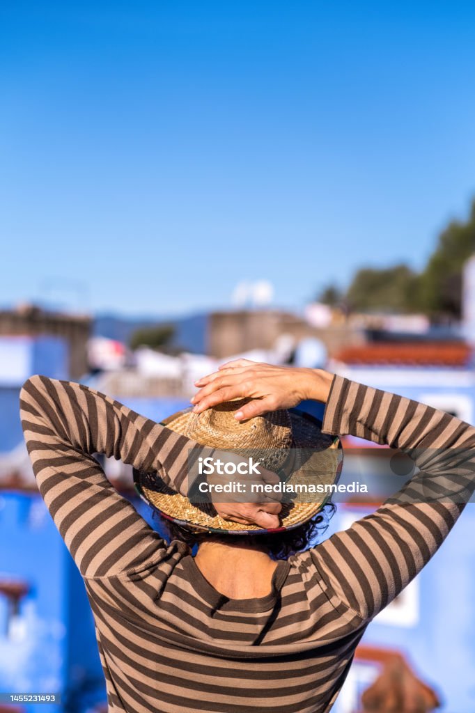 Adult woman in straw hat leaning out on the terrace of a tourist apartment at a travel destination. Rear view. Traveling connects us with other places and people. Adult Stock Photo