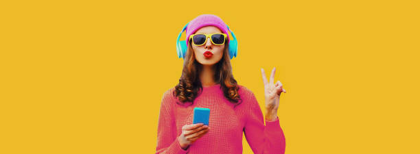 Portrait of stylish young woman in headphones listening to music with smartphone and blowing her lips on yellow background stock photo