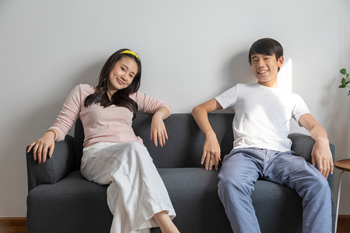 Cheerful Asian girl smiling and embracing cute boy while sitting on comfortable couch at home. Happy teenage brother and sister.