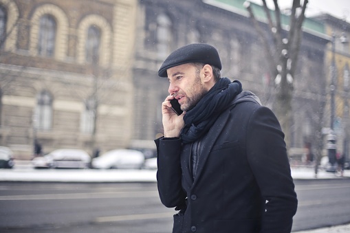 A handsome Italian man wearing a scarf and a coat standing outside in the street talking on the phone