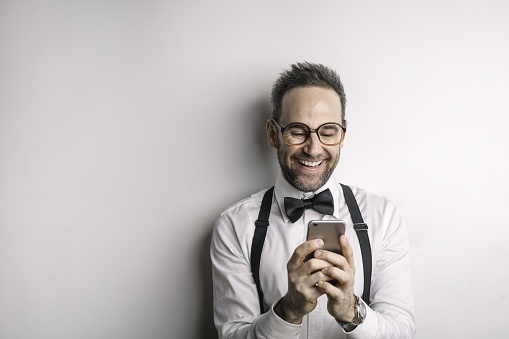 A handsome Italian man wearing a white shirt, bowtie against a white wall with his phone in hands