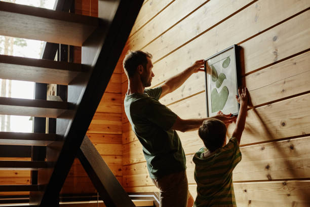 istock Side view father and son hanging picture on wall in new house, backlit 1455235530