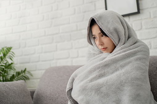Sick young asian woman sit on sofa at home wrapped covered in warm duvet or blanket, She feeling unhealthy ill.