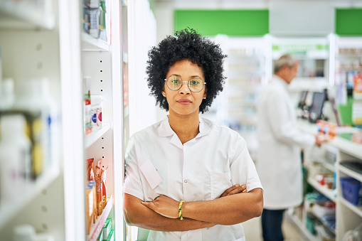 Portrait of a woman pharmacist with arms crossed leaning to the shelf in pharmacy. Female chemist in lab coat working at medical store.