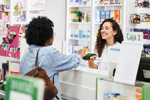 Female customer doing nfc payment with her credit card at pharmacy. Smiling cashier  holding credit card reader at drug store checkout counter.