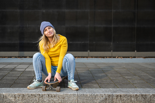 Young female teenager dressed in yellow jersey and jeans, sitting on a skateboard, looks at the camera and smiles, generation z lifestyle