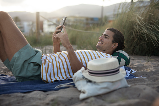 Handsome guy chilling at the beach. Young man using the phone while resting on the sandy beach.