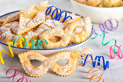 Angel wings or sfrappole or chiacchiere in shape of carnival mask. Traditional sweet crisp pastry, deep-fried and sprinkled with powdered sugar. Carnival food tradition. Decorated with paper serpentine.
