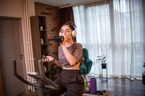 One young woman, fitness woman, rides an exercise bike in the living room, takes care of a healthy lifestyle, listens to music during training on wireless headphones.