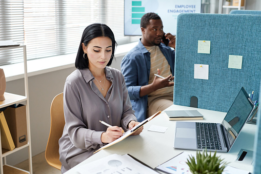 Portrait of young businesswoman writing on clipboard while working in office cubicle