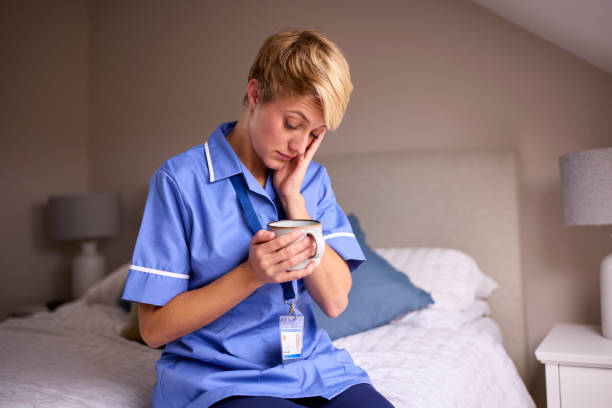 Tired Woman Wearing Nurses Or Doctors Uniform With Hot Drink Sitting On Bed At Home stock photo