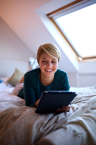 Young Woman Lying On Bed Wearing Pyjamas At Home Looking At Digital Tablet