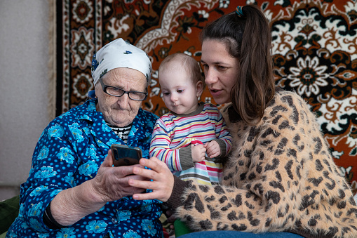 The woman with her daughter visiting her grandmother in the village. The woman with her daughter and grandmother are talking with relatives using a video call on a smartphone. Woman and grandmother are sitting on the bed in the room. The child sits between them.