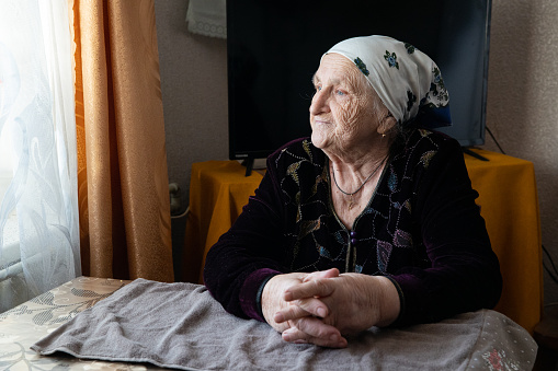 Grandmother is sitting at a table near the window in the living room at home. Senior woman looks through the window at the street.