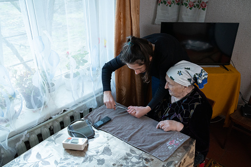The woman cuts her grandmother's nails on her hand. The woman visits a lonely grandmother to help her. Grandmother is sitting at the table near the window.