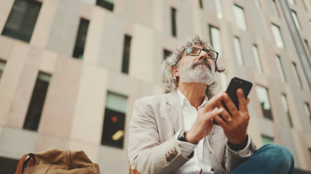 istock Mature businessman with beard in eyeglasses wearing gray jacket is using cell phone. Middle aged manager scrolling information on his smartphone while sitting outside the office 1455262598