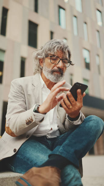 istock Mature businessman with beard in eyeglasses wearing gray jacket is using cell phone. Middle aged manager scrolling information on his smartphone while sitting outside the office 1455262615