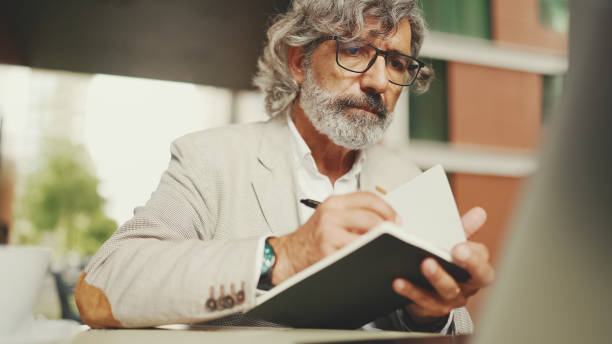 istock Mature businessman with beard in eyeglasses, user on laptop computer and taking notes, sitting in outdoor cafe. Successful man sitting at table in cafe in nature Mobile office freelancer 1455263036
