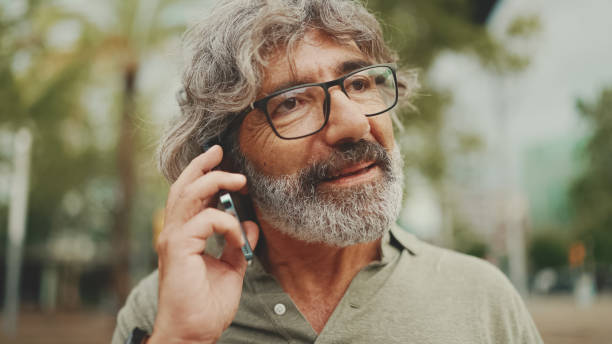 istock Friendly middle aged man with gray hair and beard wearing casual clothes using his mobile phone. Mature gentleman in eyeglasses talking on cell phone outdoors 1455263507