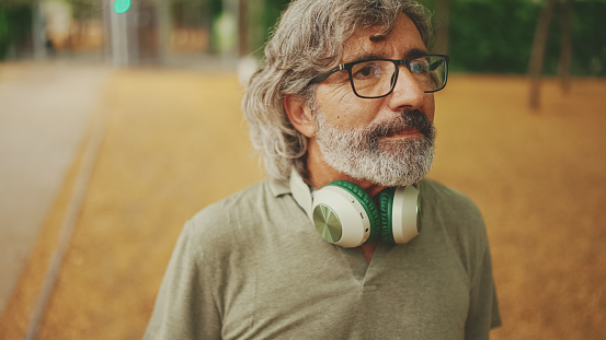Thoughtful middle-aged man with gray hair and beard wearing casual clothes with headphones around his neck is walking down the street