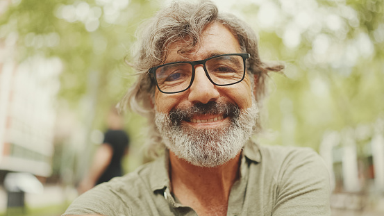 Thoughtful middle-aged man with gray hair and beard wearing casual clothes sits on bench. Mature gentleman in eyeglasses turns his head and looks into the camera