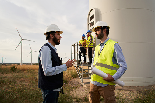 Group of engineers in hard hats talking and working with a blueprint in an agricultural field with wind turbines. Wind power station. Renewable and clean energy concept, sustainable future.